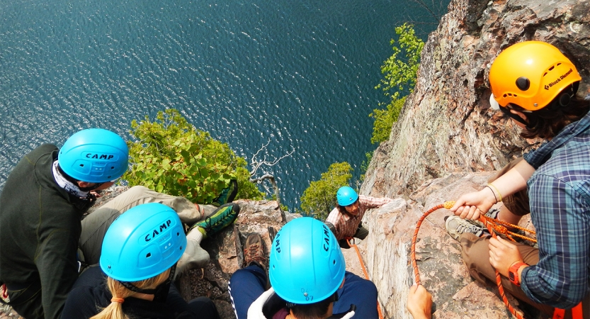 一群学生戴头盔在悬崖边上看一下面的攀岩者。下面是蓝色的水。